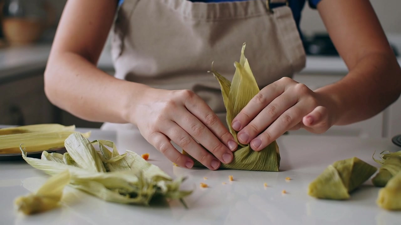 Si vede un involucro parzialmente formato, una donna con grembiule beige che piega gli involucri sul piano di lavoro della cucina, racchiudendo il ripieno.
