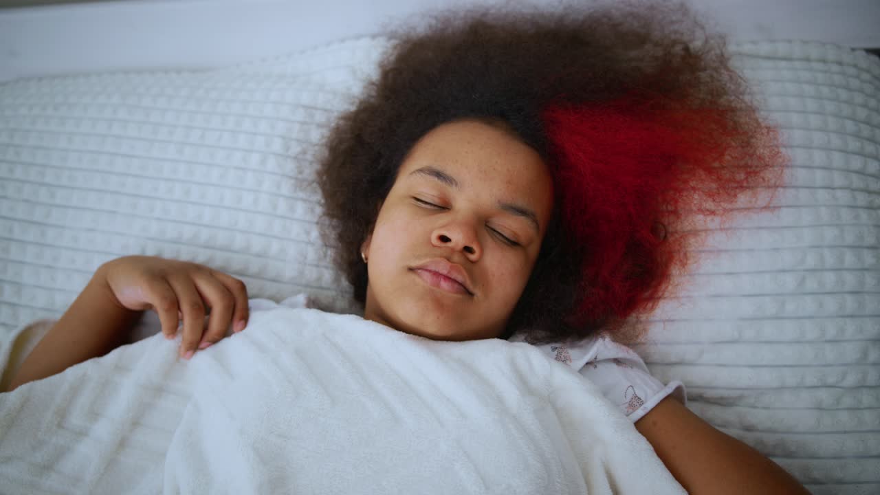 Young black woman with partially dyed red hair resting peacefully in bed, shifting positions during deep, comfortable sleep, showcasing serene nighttime relaxation