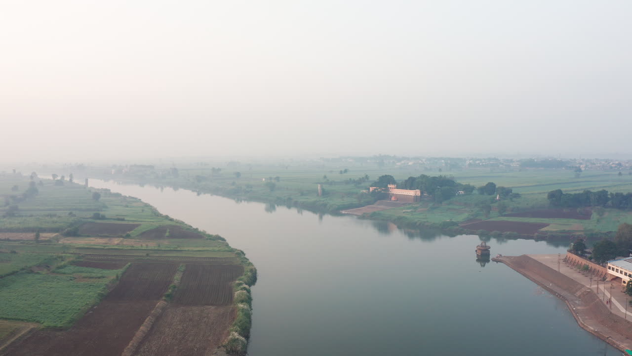 Tranquilla confluenza del fiume Krishna nel Maharashtra rurale in una mattina nebbiosa, delimitata da vivaci campi stagionali verdi.