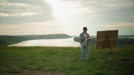 a craftsman in a black hat, checkered shirt, and jeans is engrossed in painting on a large canvas set up in a tranquil grass field by a serene lake. cloudy sky, surrounded by the beauty of nature