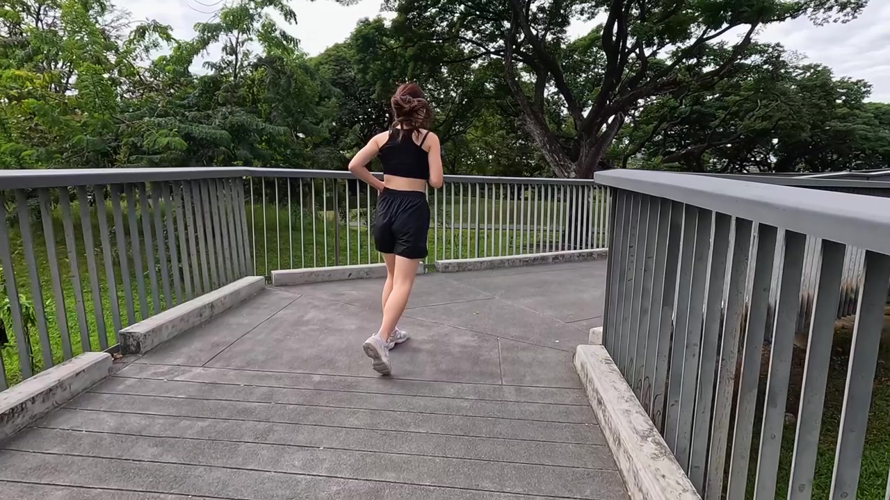 Vista posteriore di una donna in abbigliamento sportivo nero che fa jogging su un ponte in un parco lussureggiante.