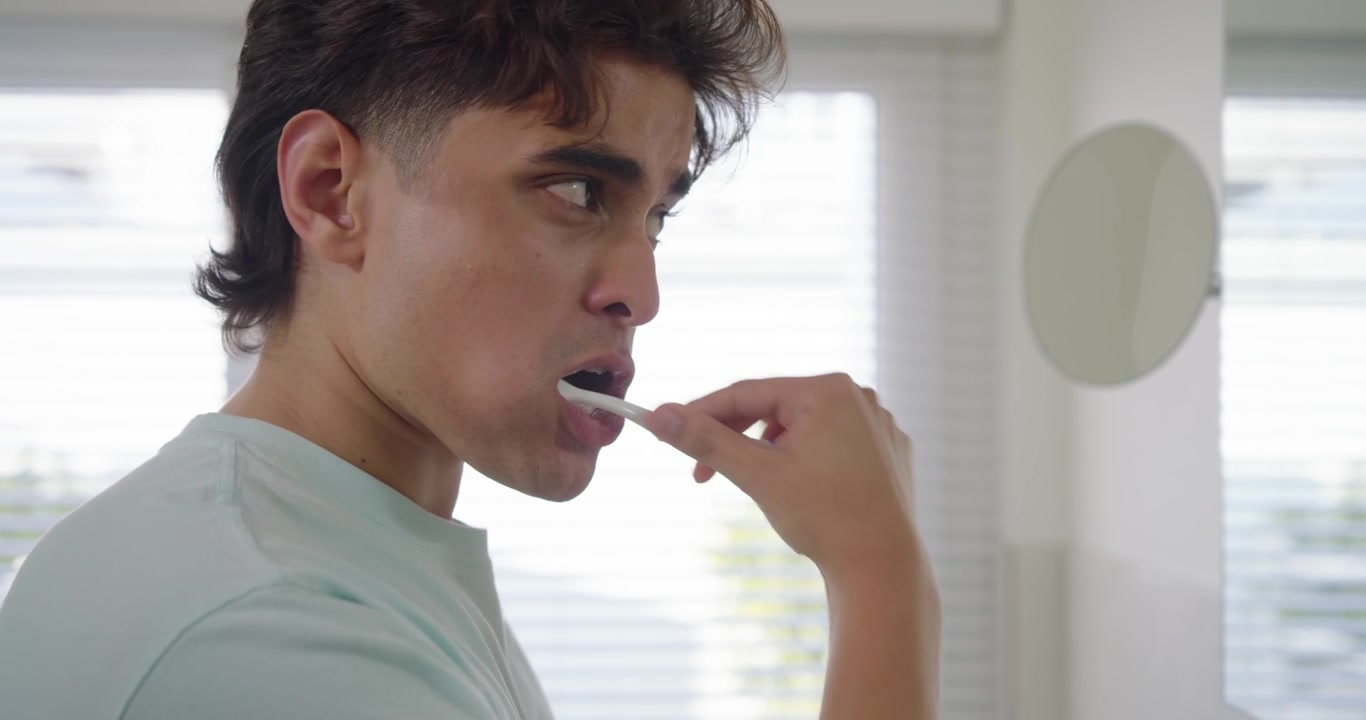 African American man brushing teeth at bathroom sink by window checking mirror for morning routine