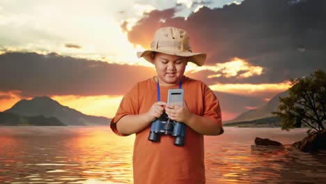 niño asiático con sombrero y binoculares mirando su teléfono inteligente y sonriendo a un lago. niño investigador examina algo, concepto de aventura de turismo de viaje