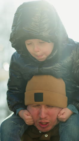 padre llevando a su hijo sonriente en sus hombros, mientras el niño juega con su barbilla, haciendo que el rostro del padre se gire, rojo por el peso del niño, con suaves rayos de luz por detrás