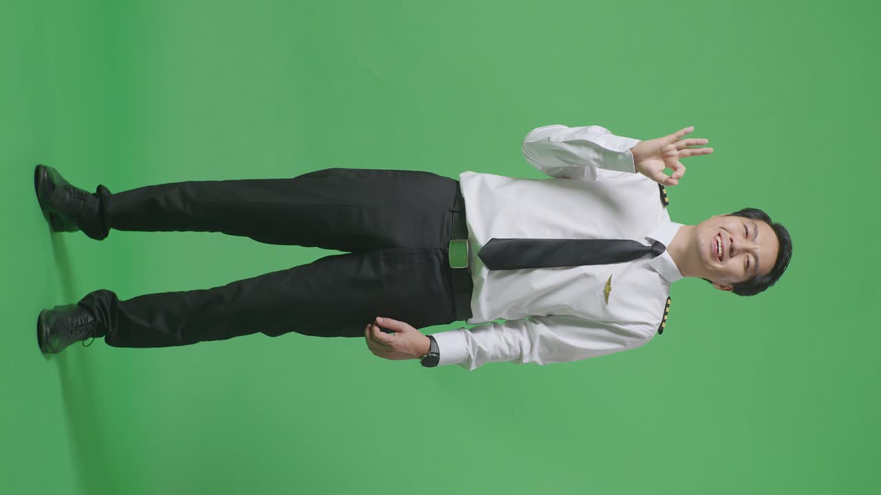 아시아 남성 조종사가 미소 짓고 녹색 스크린 배경 스튜디오에 서있는 동안 카메라에 은 몸짓을 보여주고 있습니다.