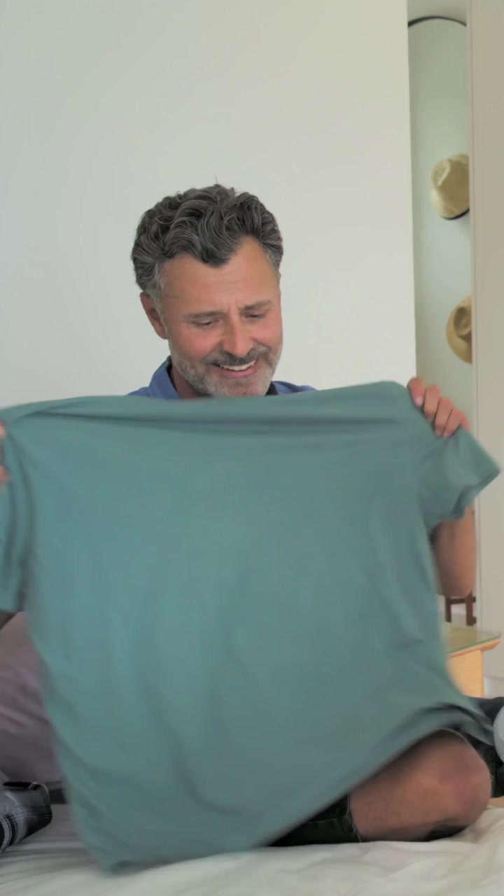 Vertical video: Folding man in blue folding teal shirt on bed, on partner entry twirling by basket