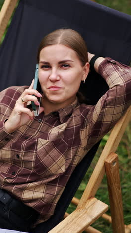 relaxed happy caucasian lady speaks on the phone lying in the garden chair. free time freelancer having during remote work. outdoors setting. vertical video