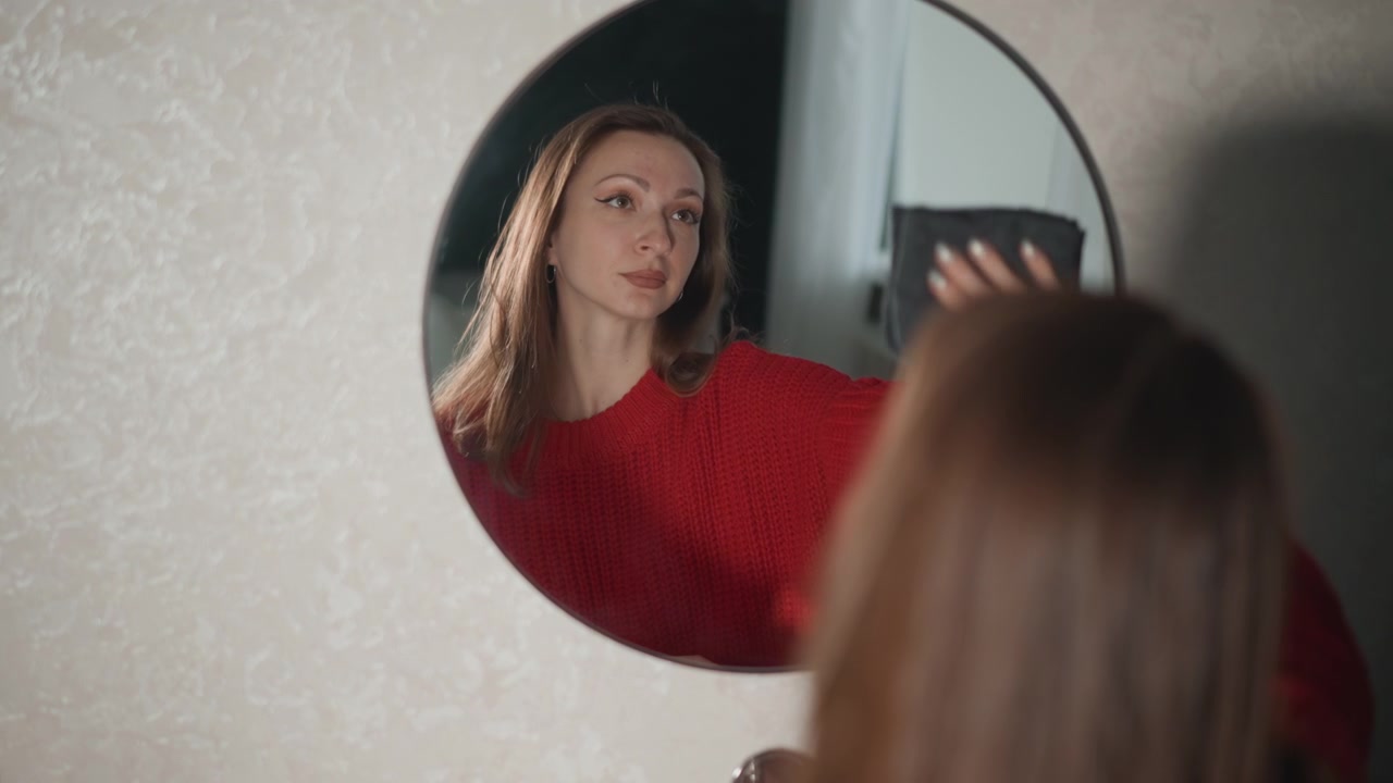 Donna con maglione di lana rosso alza lo smartphone accanto a uno specchio rotondo a parete, controllando il suo riflesso con uno sguardo concentrato, una leggera inclinazione della testa, una postura rilassata, una luce interna soffusa e un ambiente domestico pulito.