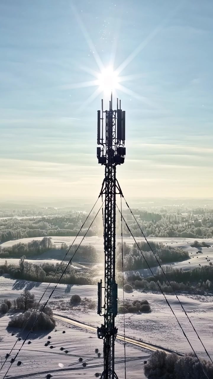 A vertical shot of a communication tower with the sun above in a snowy winter landscape.