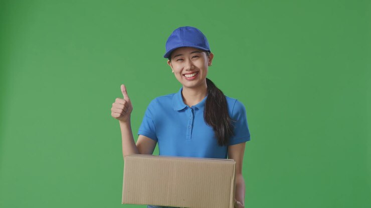 Asian Female Courier In Blue Uniform Showing Thumbs Up Gesture And Smiling While Delivering A Carton On Green Screen Background In The Studio
