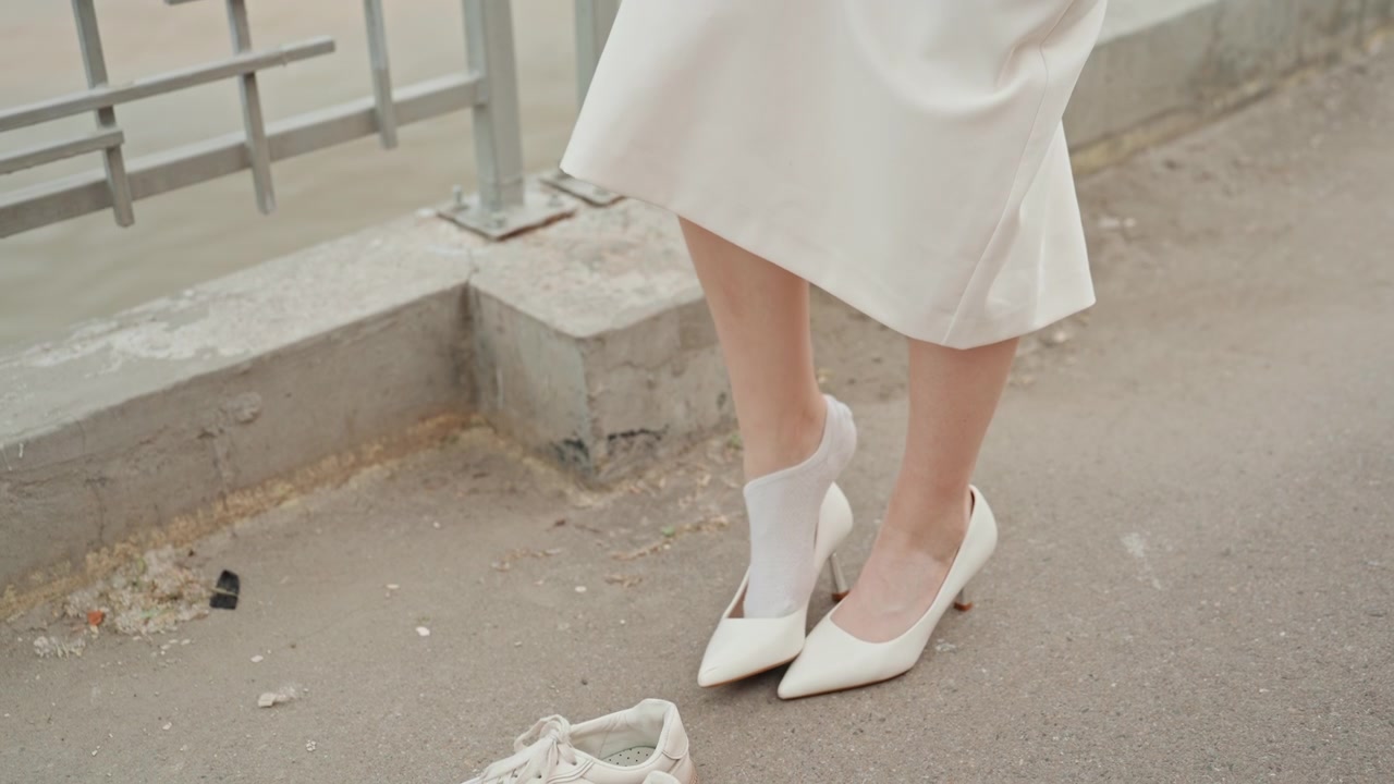 Mulher tira sapatos brancos de salto alto e calça meias em um calçadão à beira do rio. A mulher levanta o vestido para ajustar a meia, com tênis casuais por perto no pavimento de concreto perto de grades de metal.