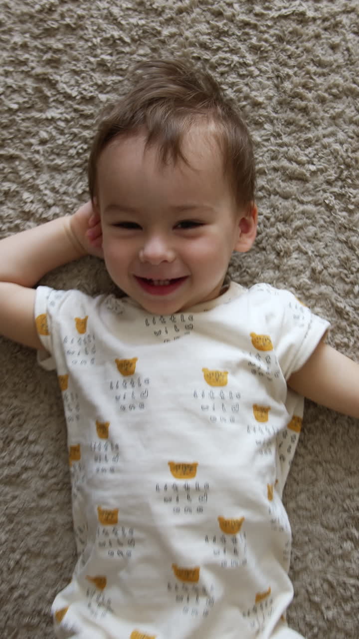 Camera approaches a beautiful Caucasian baby boy lying on the carpet. Kid smiles to camera and turns on right side. Top view. Vertical video
