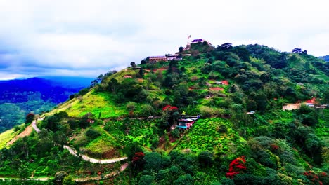 a lush sri lankan hill rises with a buddhist temple on top, winding dirt paths, scattered village houses, and a breathtaking tropical mountain backdrop under a moody, cloudy sky.