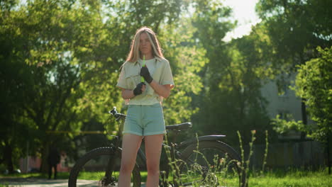 young lady stands in bright sunlight, holding green air pump near face beside bicycle, looking focused as her hair flutters, background features blurred trees, building, and figure walking