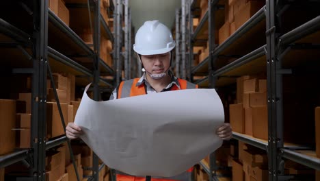 asian male engineer with safety helmet looking at blueprint in his hands and looking around while standing in the warehouse with shelves full of delivery goods