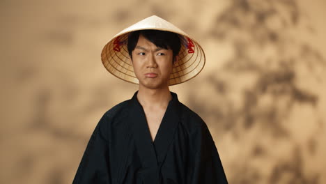 Young-Asian-man-in-black-traditional-costume-and-conus-hat-looking-at-camera-with-angry-face