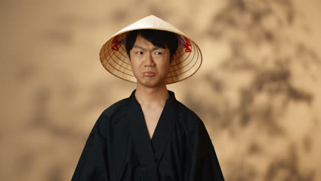 young asian man in black traditional costume and conus hat looking at camera with angry face