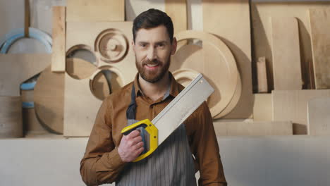 portrait of caucasian bearded man in helmet and holding saw smiling at camera in carpentry workshop