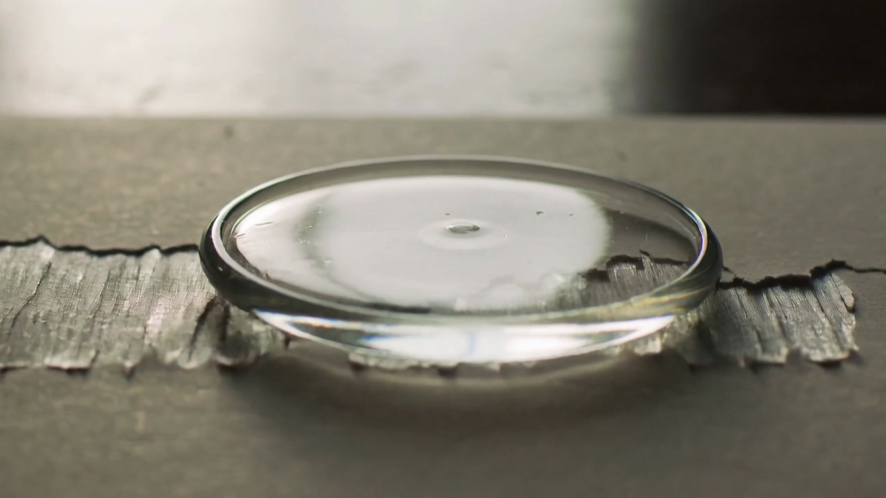 Shifting convex droplet touching torn cardboard tear, sliding left and relaxing into fibers