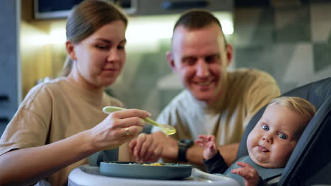 attractive caucasian woman feeding her baby boy. dad sitting behind watches and smiles. blurred backdrop.