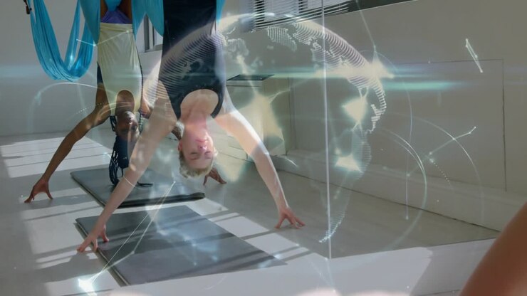 two women performing inverted pose with blue hammock in yoga studio, showing digital health overlay