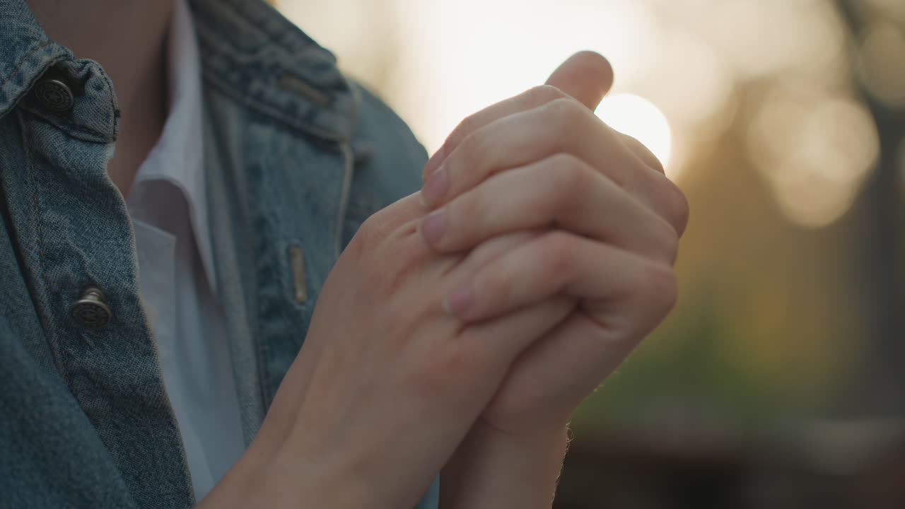 Sunlit hands clasped, young white woman reflecting on family future, gentle smile, denim jacket, golden hour park, intimate closeup on fingers, daughter role, tender contemplative mood