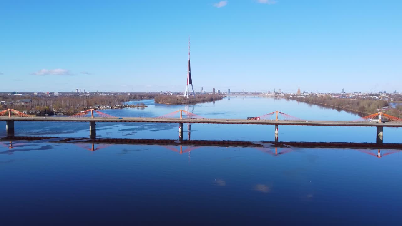 Drone aerial view of bridge crossing the calm Daugava River in Riga, Latvia with mirror reflection on the water and distant Riga TV Tower