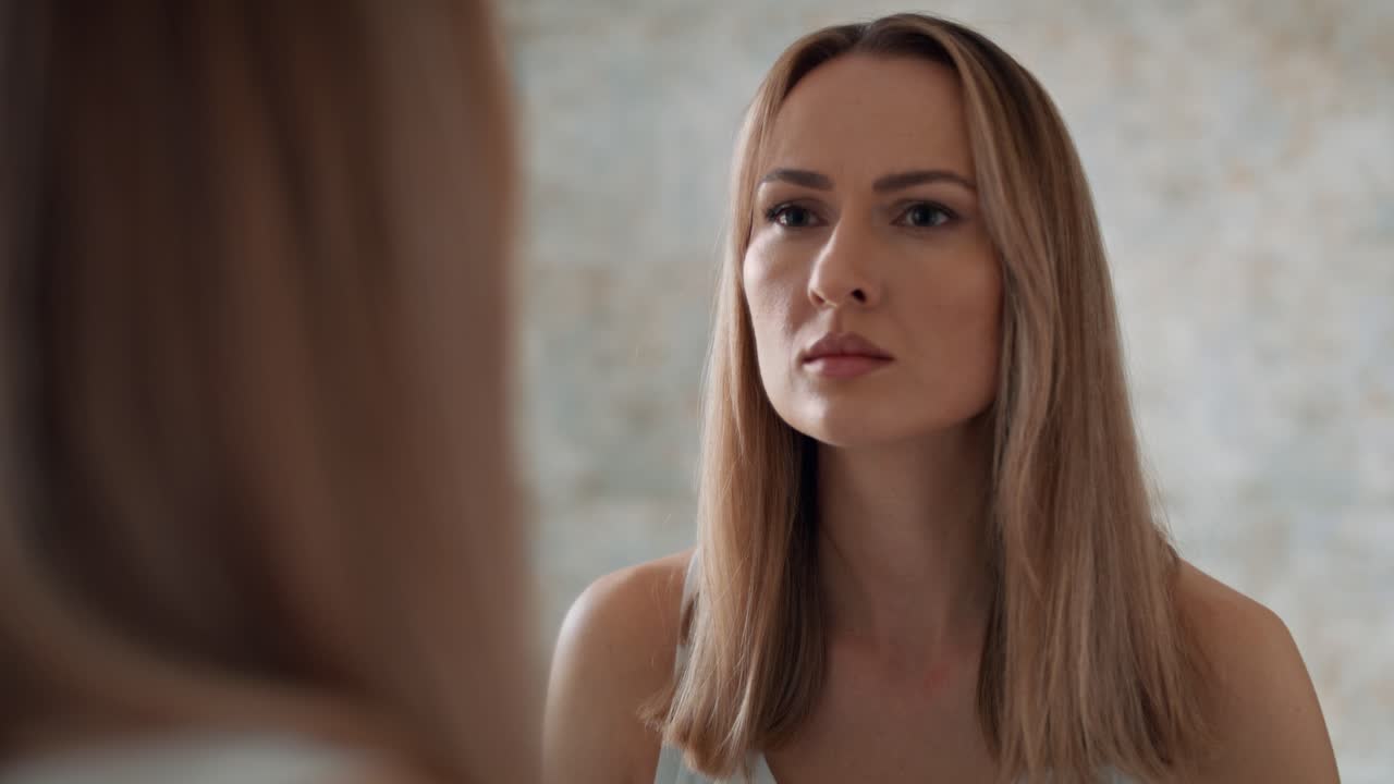 Serieuze vrouw staat voor de badkamerspiegel en kijkt naar zichzelf.