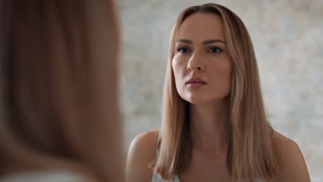 Serious-woman-standing-in-front-of-bathroom-mirror-and-looking-at-herself.