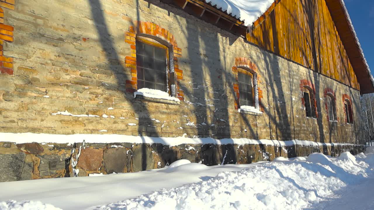 Old limestone stone or rock building with red bricks around windows. The architecture is old and beautiful. Sun is shining on the building and white fluffy dense snow around it. Roof covered in snow.