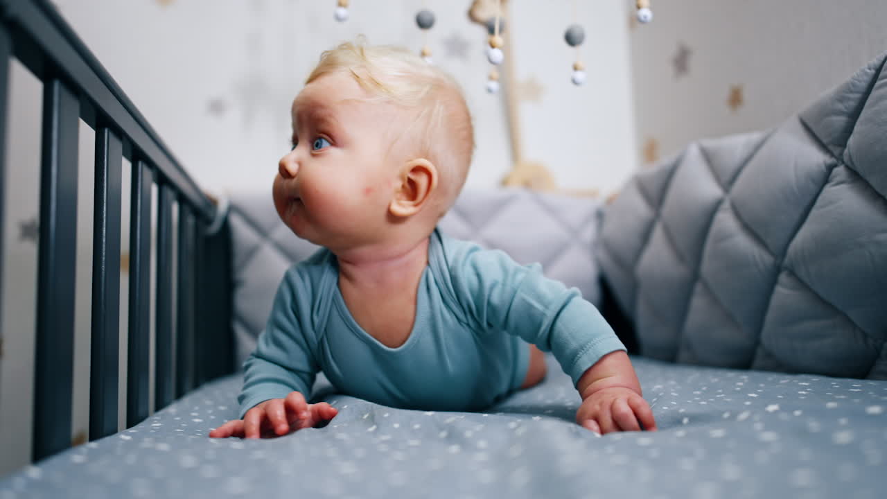 Tiny blue-eyed Caucasian infant in his crib. Adorable baby boy trying to look above the handrail of a cot.