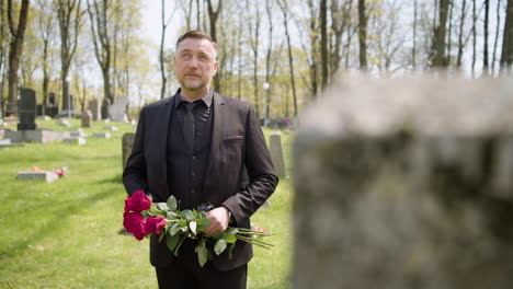 sad man in black suit standing in a graveyard holding red roses while looking at the sky and at tombstones sorrowfully