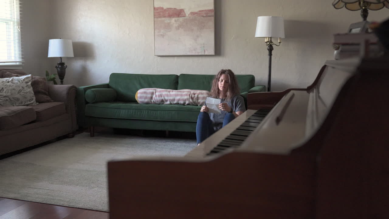 A woman sits on the ground, leaning against a green couch as she reads a love letter. The camera slides from right to left behind a piano, gradually revealing more of her serene expression.