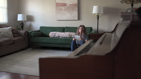 a woman sits on the ground, leaning against a green couch as she reads a love letter. the camera slides from right to left behind a piano, gradually revealing more of her serene expression.