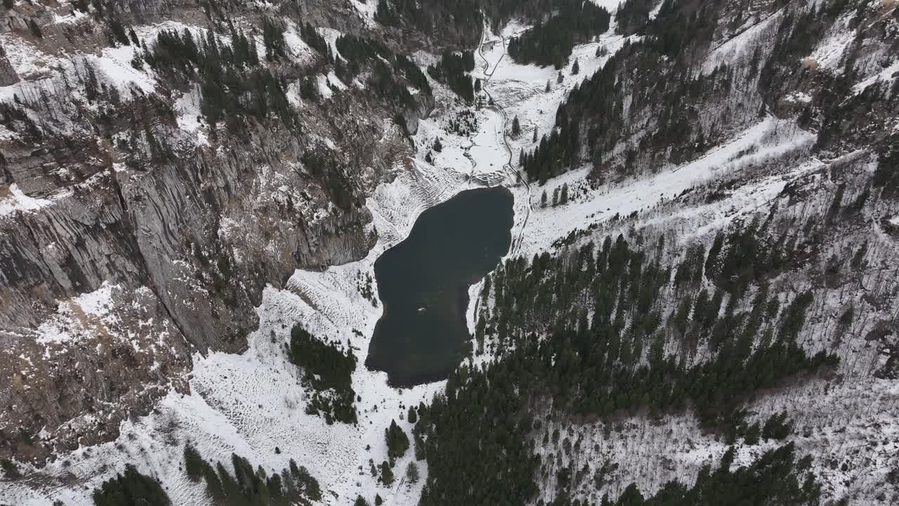 vista de pájaro de un pequeño lago entre valles de montaña cubiertos de árboles de hoja perenne y nieve