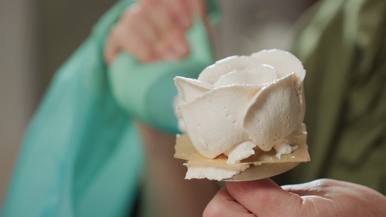 Close up of caterer using green piping bag to carefully coat icing in petal-like shape on rotating baking tool, highlighting precision dessert decorating in soft-lit kitchen setting