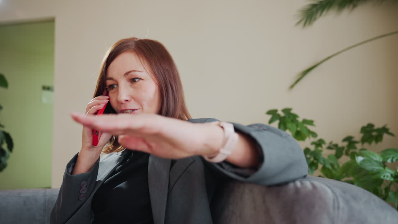 Focused businesswoman in gray blazer speaking on phone while gesturing during intense conversation, seated in cozy office environment with lush indoor plants and natural lighting