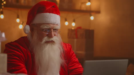 santa claus real usando nueva tecnología para la comunicación con los niños que reciben correo o lista de deseos. alegre trabajando en la computadora portátil y sonriendo mientras está sentado en su silla con chimenea y árbol de navidad. imágenes de alta calidad 4k