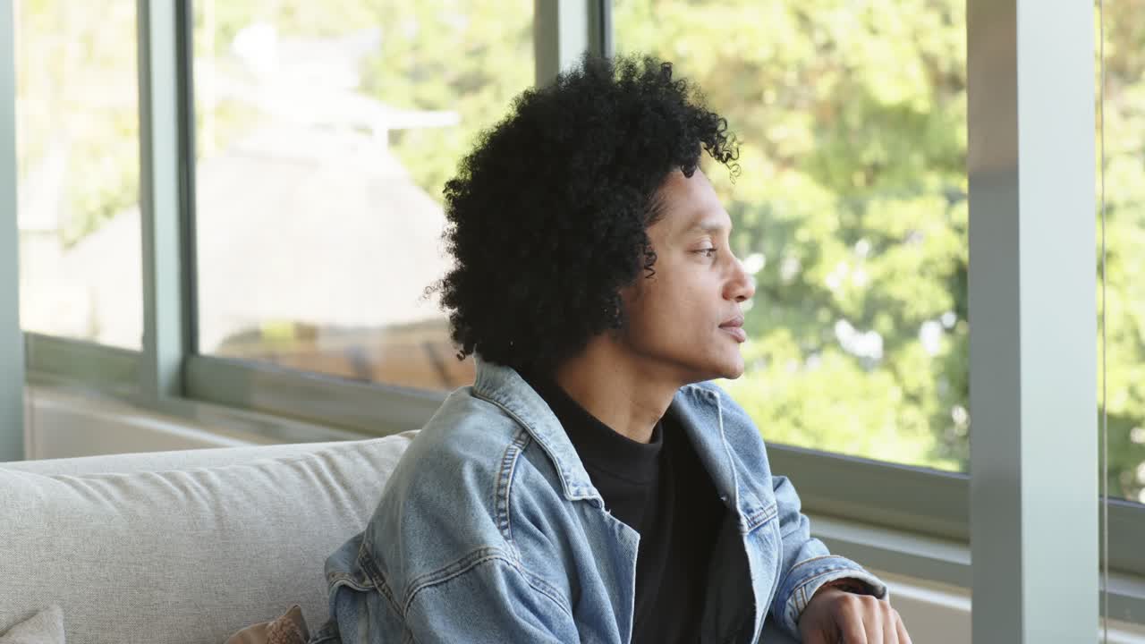 Uomo afroamericano che si appoggia al divano vicino alla finestra, cambia posizione e appoggia il mento