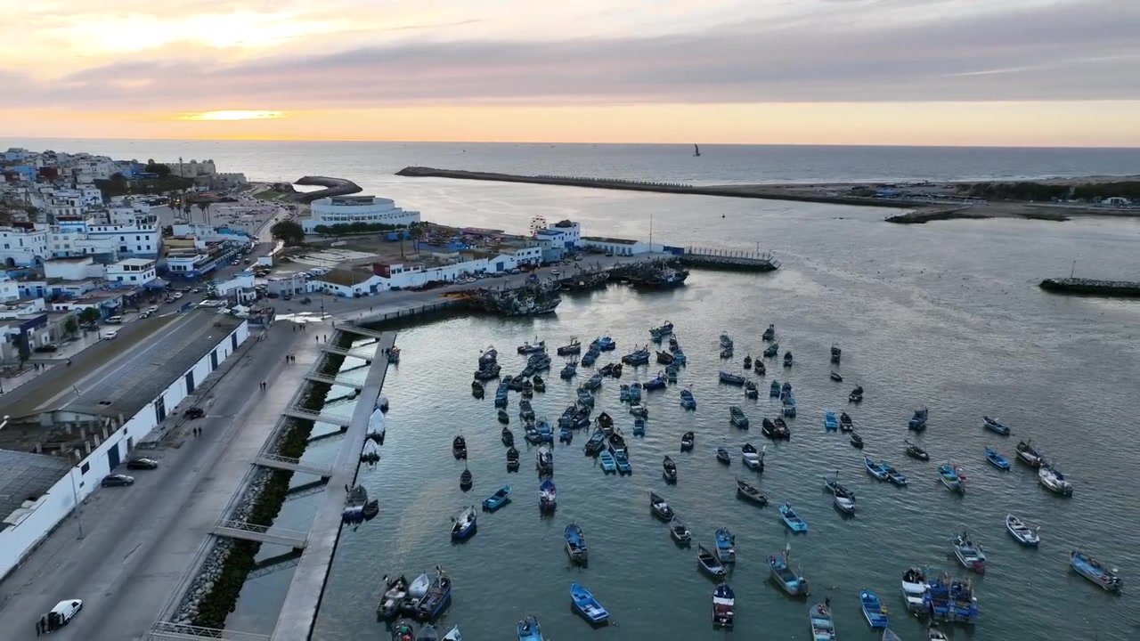 Ampia vista aerea di una piccola flotta di pescherecci all'interno del porto di Asilah al tramonto, con acqua calma e protetta, frangiflutti, città costiera e la morbida luce serale atlantica.