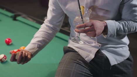 close-up of person in white shirt and grey trousers holding lemon drink with black straw, sitting on green pool table with colorful billiard balls. stylish indoor setting with soft lighting