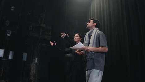 confident male brunette stage director in a scarf and gray t-shirt walks along the stage with his student actress and expressively plays the role reading words on stage in the theater