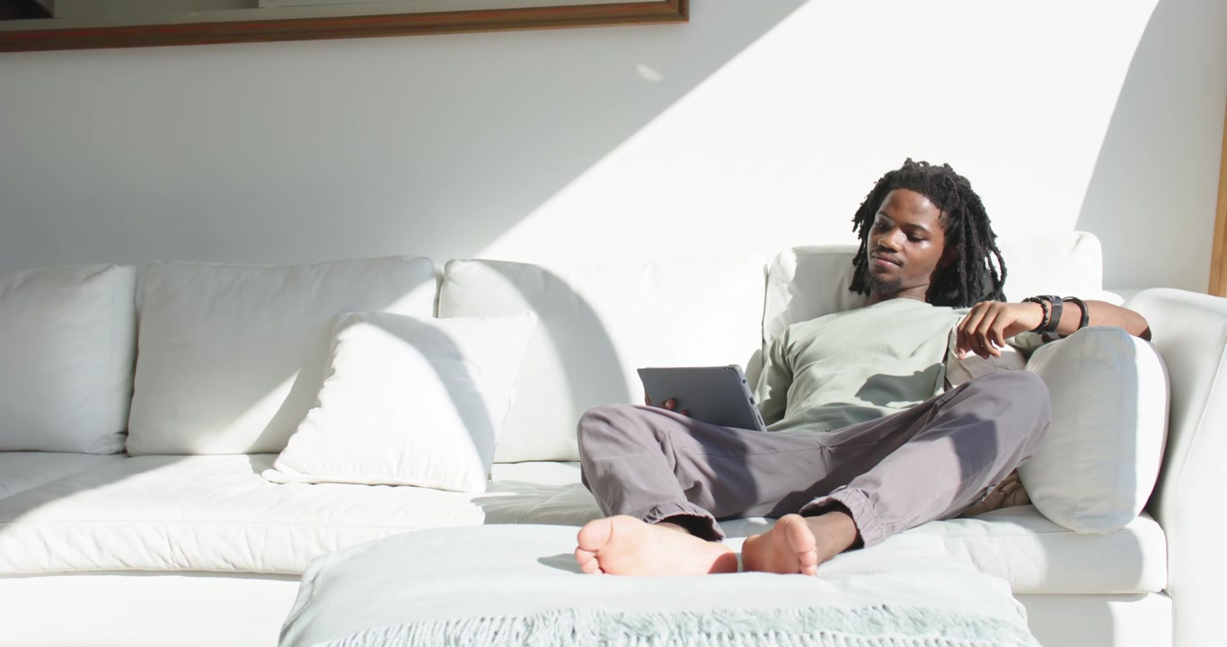 Afroamerikansk mand læner sig tilbage på højre hvide sofa, flytter vægten og læser på en tablet, med plads til tekst.