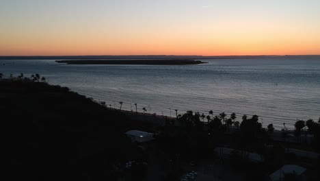 Flying-out-over-a-dark-La-Paz,-Mexico-toward-a-calm-Pacific-ocean-as-the-sunset-dips-below-the-horizon