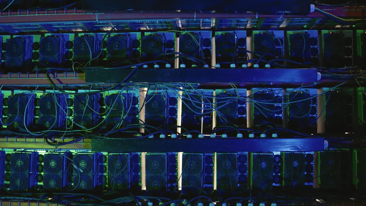 Horizontal shelves of ASIC bitcoin mining units with power and data cables, blue lighting. Tilt-down view reveals multiple stacked rows