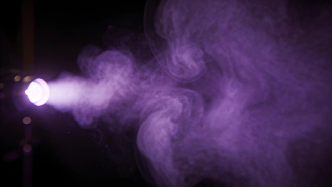 Emettitore rotondo che proietta una nebbia illuminata di viola attraverso uno studio buio, formando particelle e vortici.
