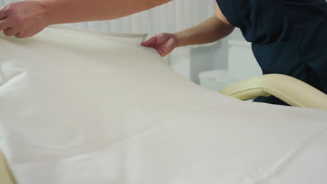 beauty technician in navy uniform spreads fresh linen over cream massage table in bright white treatment room, ensuring smooth, clean surface and professional setup for next spa and wellness session