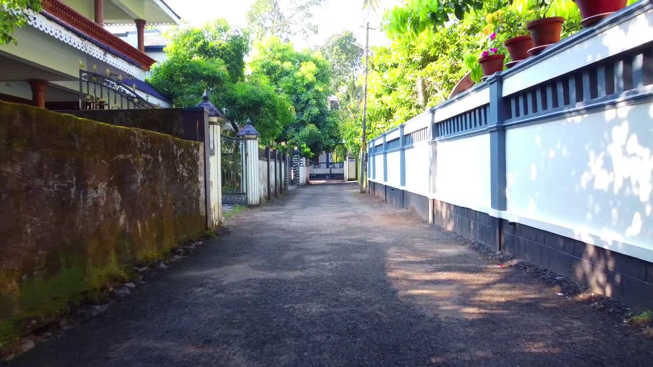 A view of a quiet, narrow residential street in Kerala, India, lined with traditional homes and lush green foliage, offering a glimpse into local, everyday life