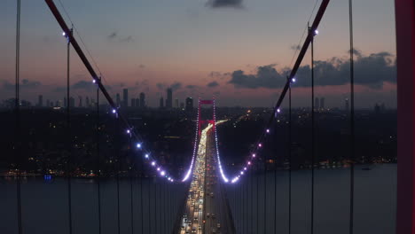 Epic-Dolly-forward-through-Istanbul-Bosphorus-Bridge-Arch-Illuminated-in-Red-light-at-Night-with-Car-traffic,-Aerial-Drone