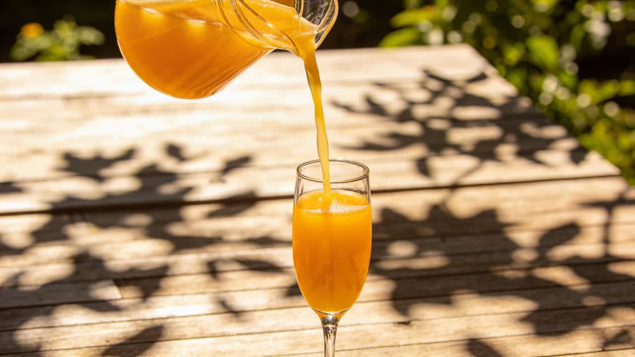 Mano fuori campo che inclina una brocca di vetro e riempie un calice a stelo su un patio in legno, con ombre di foglie.